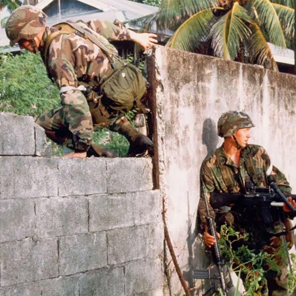 Two soldiers in camouflage uniforms climb over and guard a concrete wall in a tropical setting.
