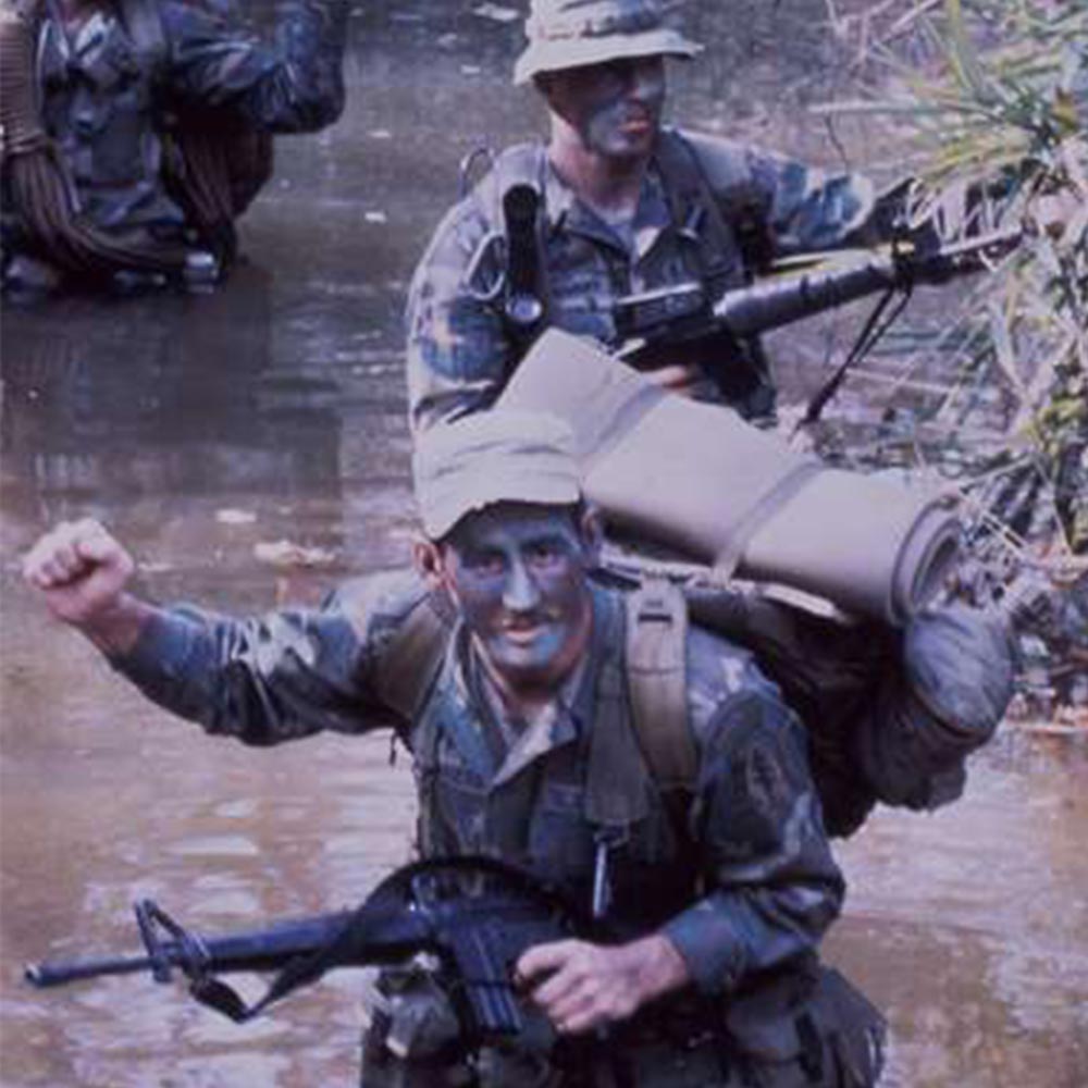 Two soldiers with face paint wade through muddy water, carrying gear and rifles, one raising a fist.