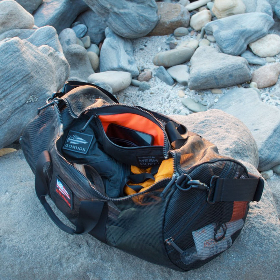 A Mesh Gym Bag with black mesh and orange lining sits open on rocky ground, revealing gear and clothing inside.