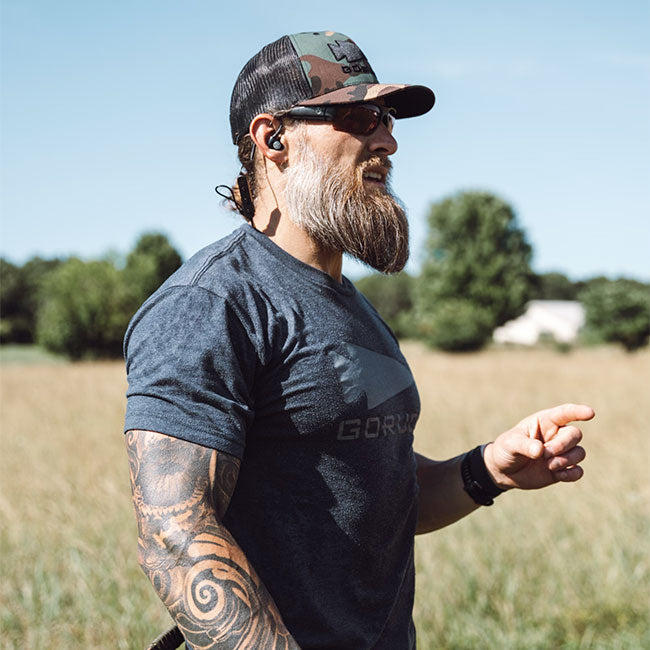 A bearded man with tattoos, sunglasses, and cap stands outdoors in a grassy field, pointing to the right.