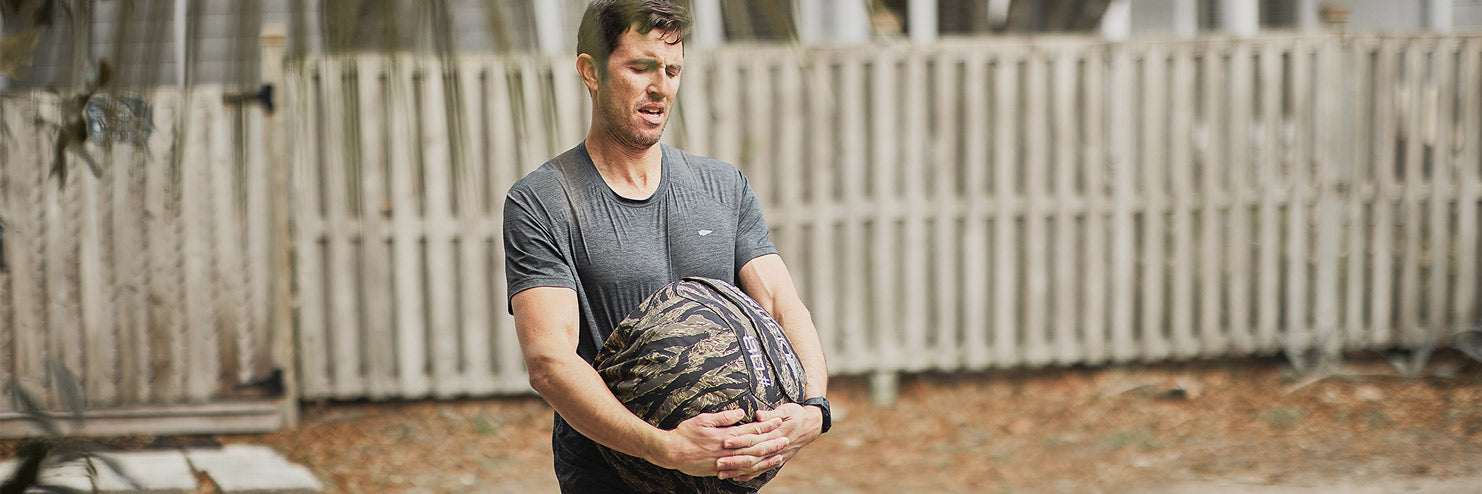 Man outdoors, straining as he lifts a heavy sandbag, with a wooden fence in the background.