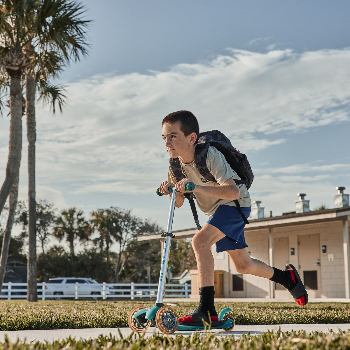 Boy in shorts and a backpack rides a scooter on a sidewalk with palm trees and buildings in the background.