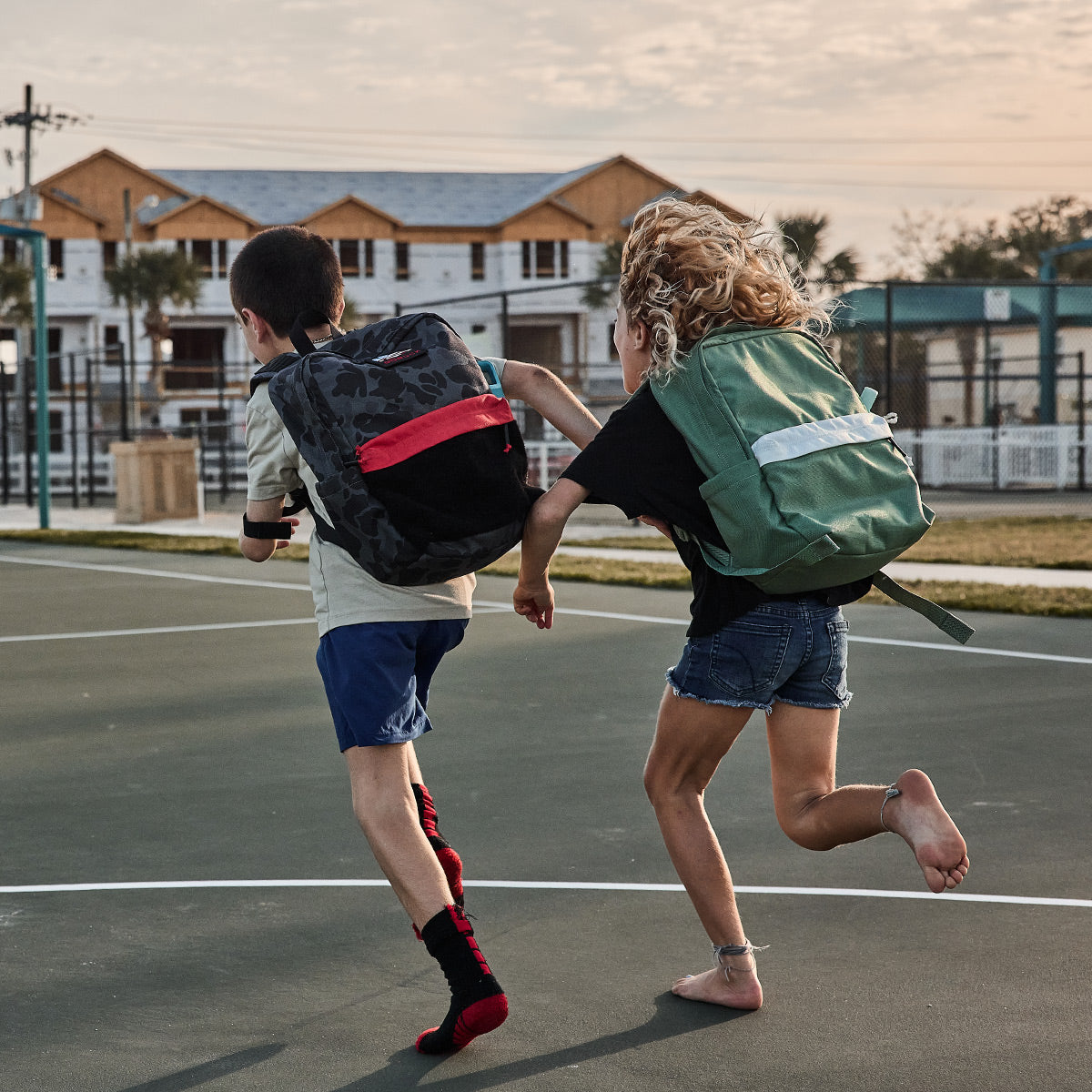 Two kids with backpacks run playfully on an outdoor basketball court near houses under construction.