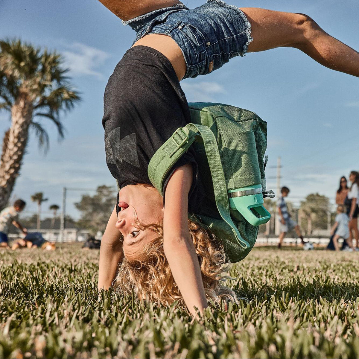 Young girl doing a handstand on grass, wearing a backpack, with palm trees and blue sky in the background.