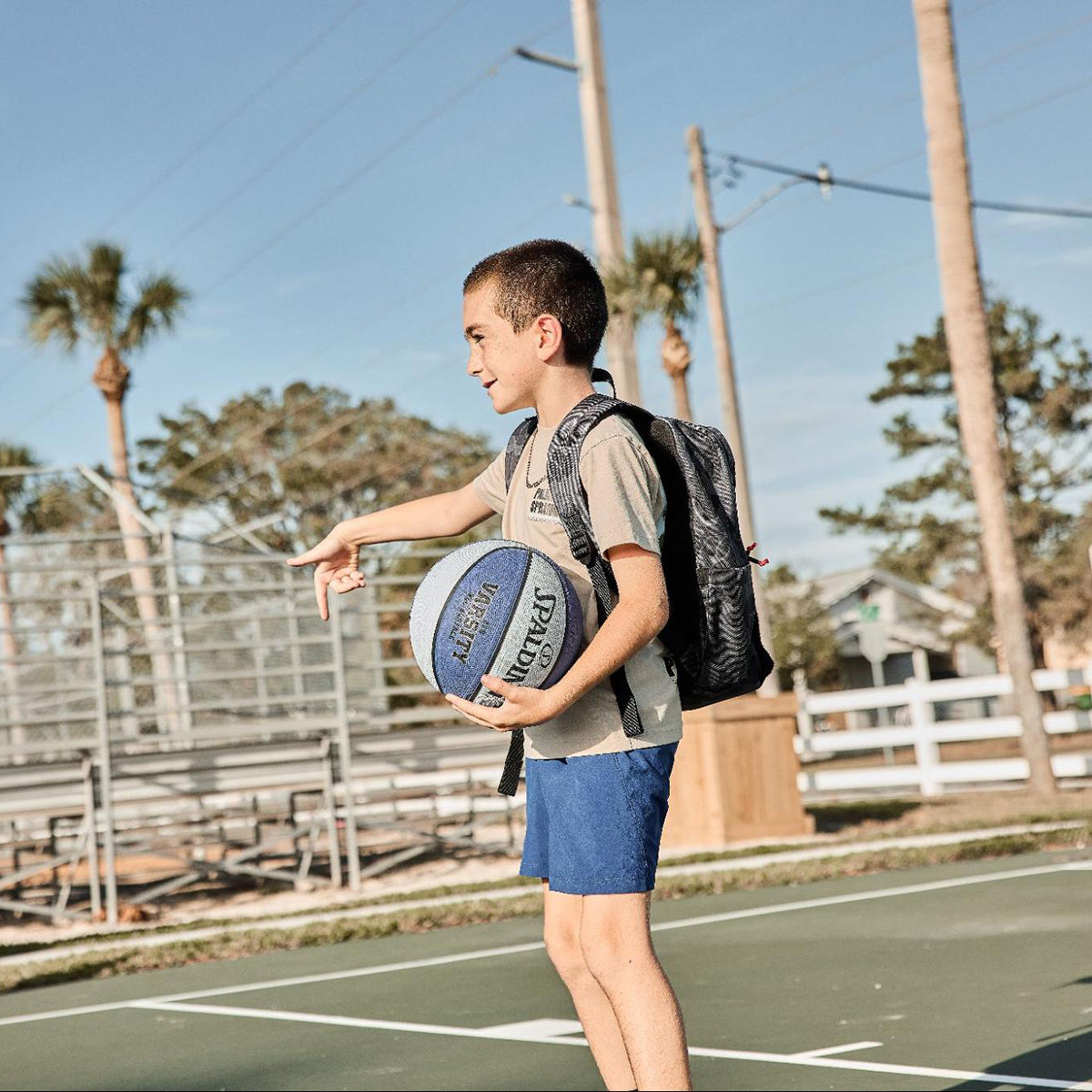 A boy in blue shorts holds a basketball and points while standing on an outdoor court with palm trees.