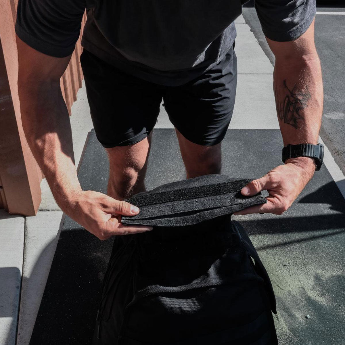 A person in shorts loads a rectangular object into a black backpack on a sidewalk.