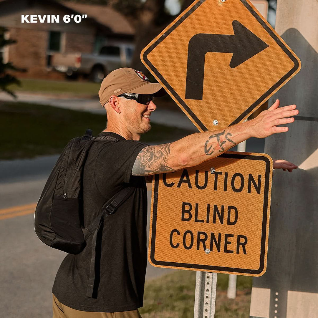 A man stands by a "Caution Blind Corner" sign, his arm level with the sign while wearing the Basic Rucker® weighted rucksack; text reads "KEVIN 6'0".