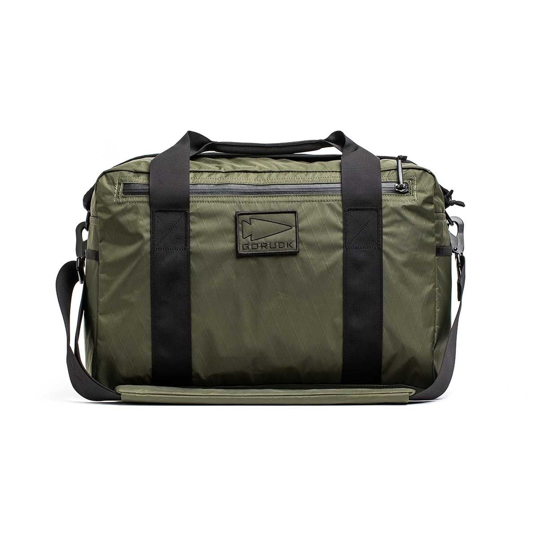 The Kit Bag - X-PAC in olive green features black straps, a front logo patch, and is made from durable X-PAC VX21 fabric, displayed against a white background.