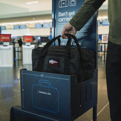 A traveler places a sleek black Kit Bag - Dyneema, made by GORUCK, into a personal item size checker at the airport. Next to it is a sign outlining luggage dimension guidelines, while in the background, check-in kiosks and counters are bustling with travelers.