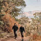 Two people hiking on a dirt trail surrounded by autumn trees with mountains in the background.