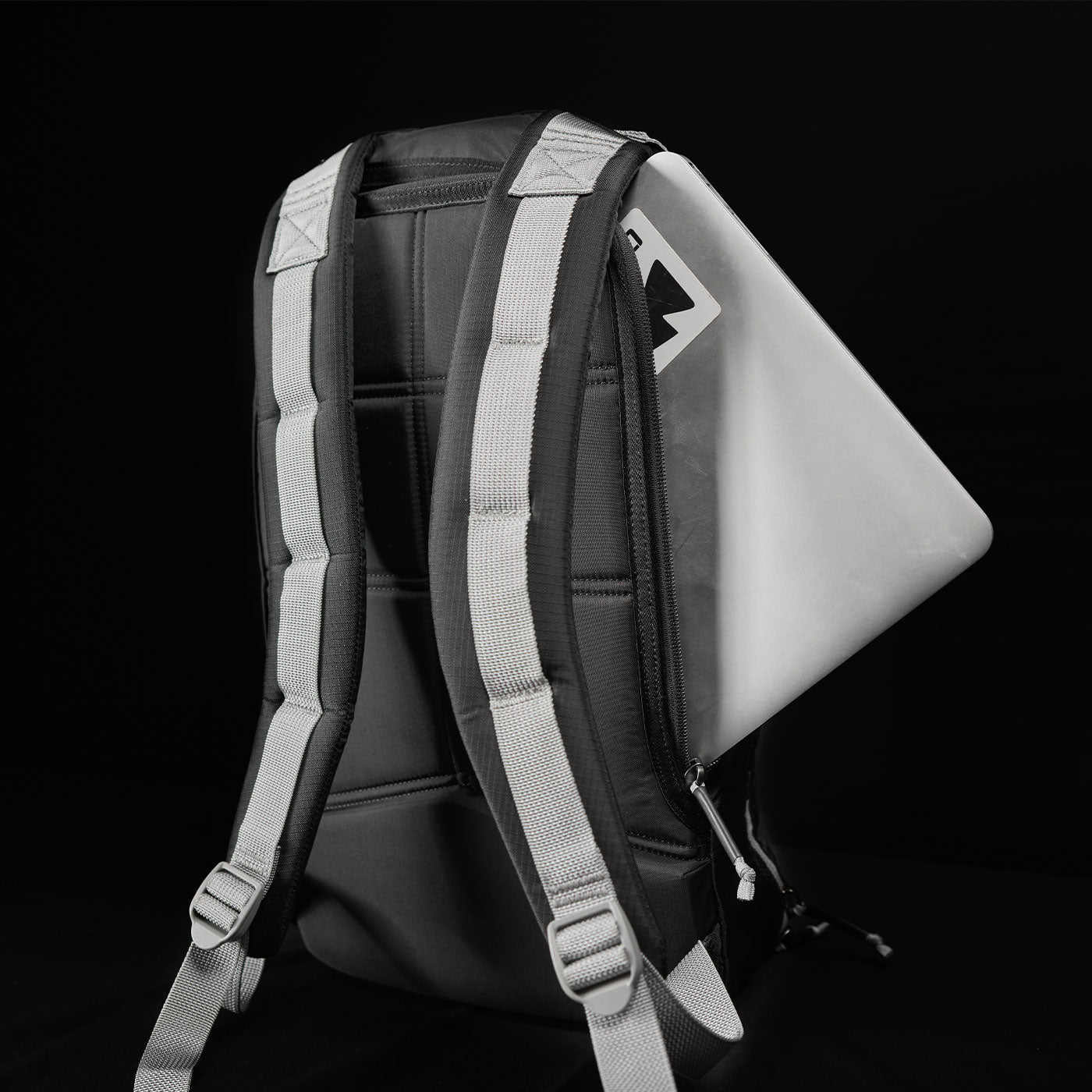 A laptop partially tucked into the back pocket of a black backpack with gray straps, against a black background.