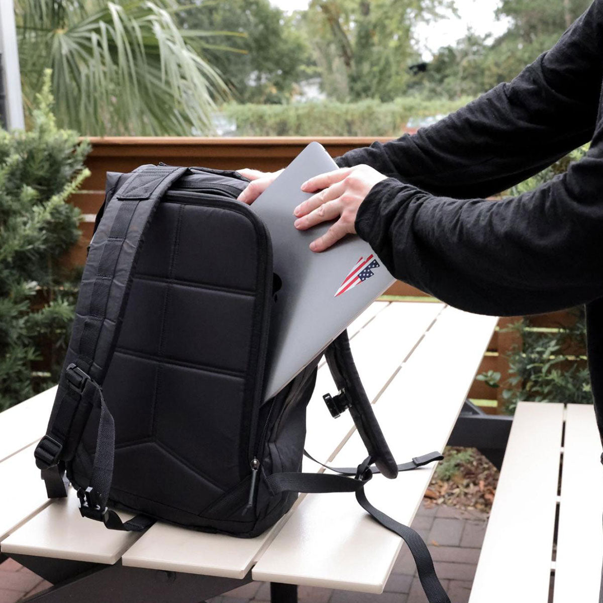 Person placing a laptop with stickers into a black backpack on a picnic table outdoors.