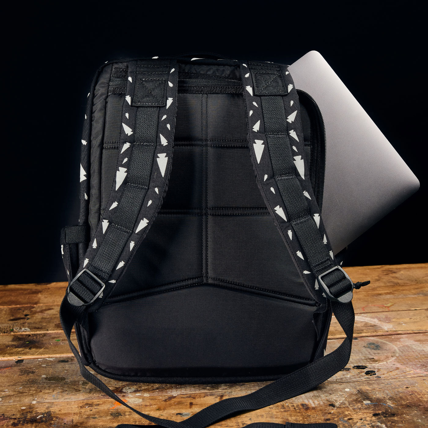 A black backpack with patterned straps sits on a wooden table, with a laptop partially inside.