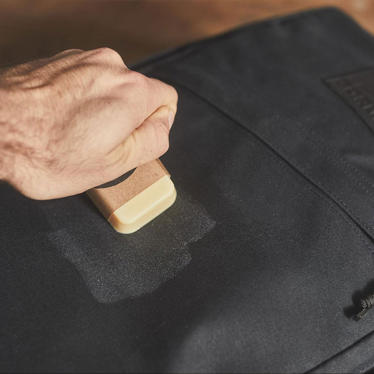 A hand rubs a bar of wax on a black fabric bag to waterproof it.