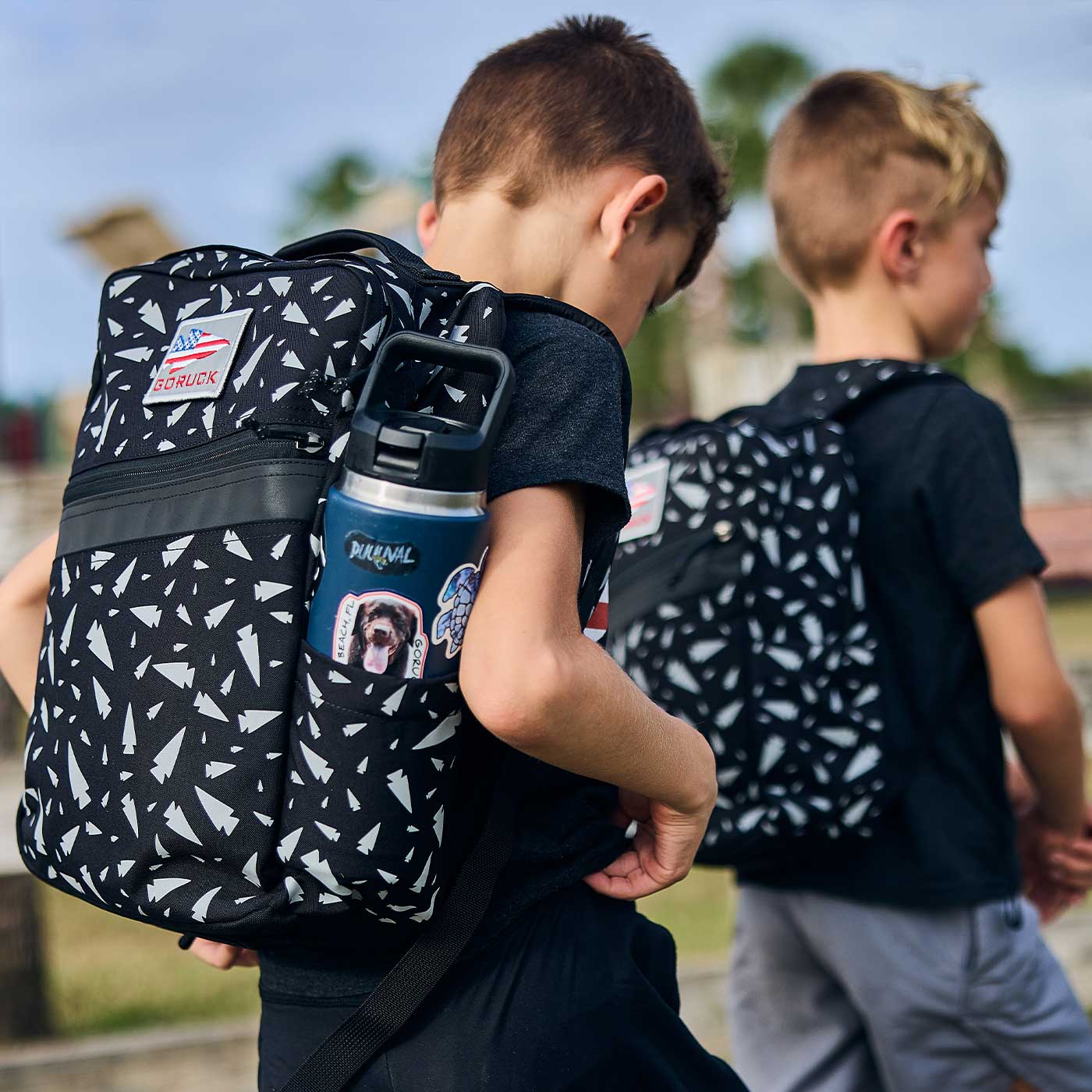 Two young boys with black patterned backpacks walk outdoors; one backpack has a water bottle and stickers.