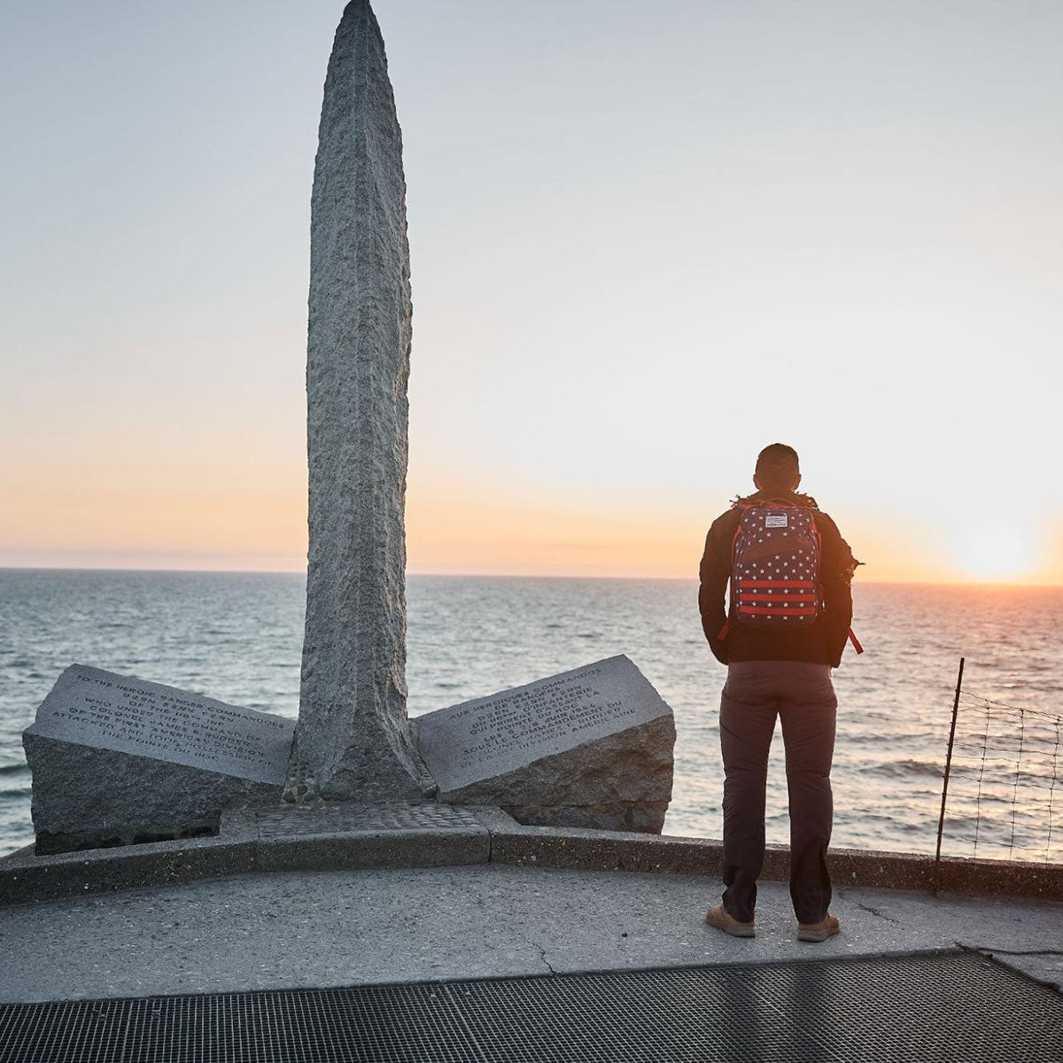 Person with a backpack stands by a stone monument by the sea at sunset, looking toward the horizon.