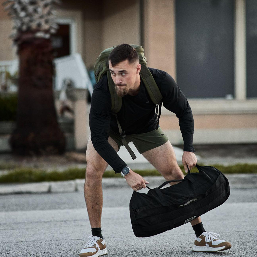 A man in athletic wear lifts a black duffel bag outdoors, wearing a backpack and sneakers.