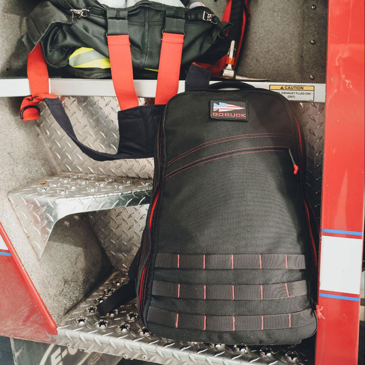 A black backpack with red stitching sits on a metal shelf next to a harness with red straps.