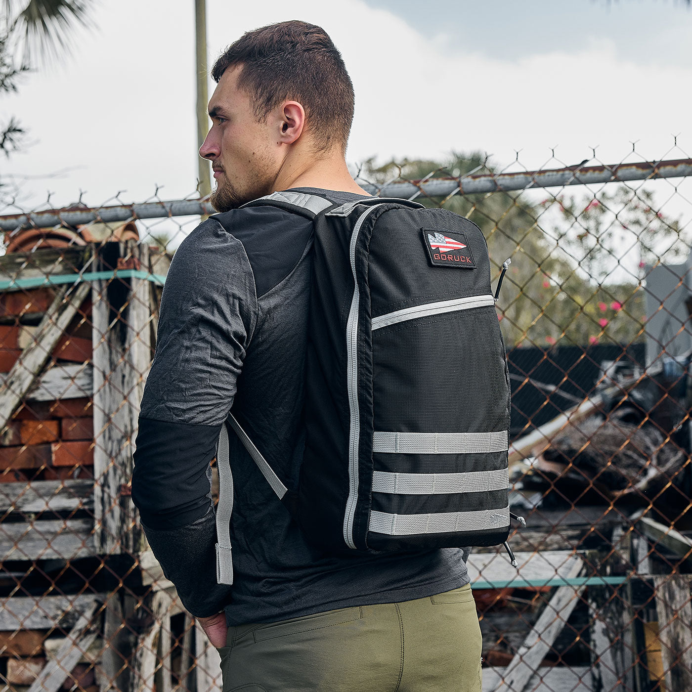 Man wearing a black backpack with reflective stripes, standing near a chain-link fence and stacked bricks.