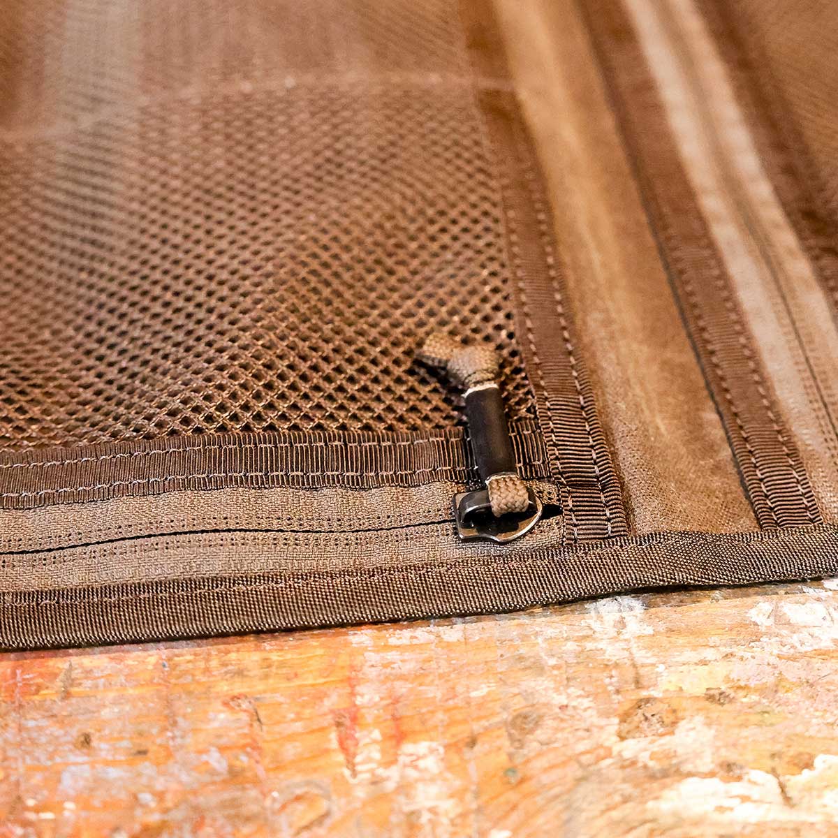 Close-up of a brown mesh zippered pouch on a wooden surface, showing the zipper pull and stitching.