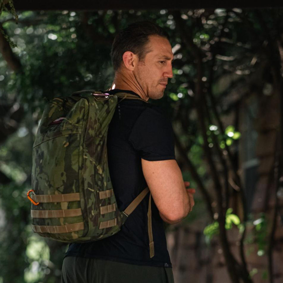 Man wearing a camouflage backpack and black shirt stands outdoors, looking over his shoulder.