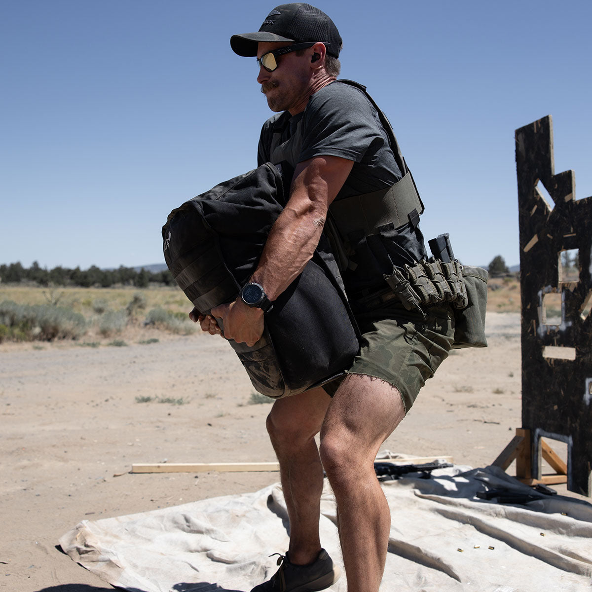 Man in tactical gear lifts a heavy sandbag outdoors on a sunny day, with dry grass and shrubs in the background.