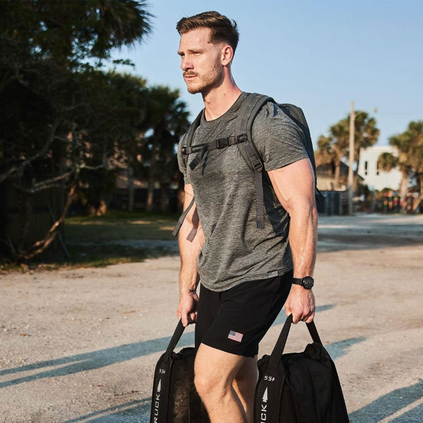 Man in athletic wear carrying two heavy bags outdoors, wearing a backpack, with trees in the background.