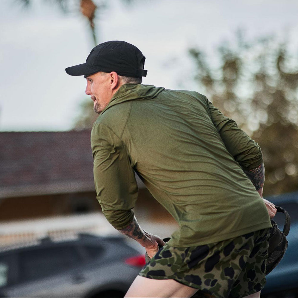 Man in green jacket and camo shorts running outdoors, wearing a black cap, with trees and cars in the background.