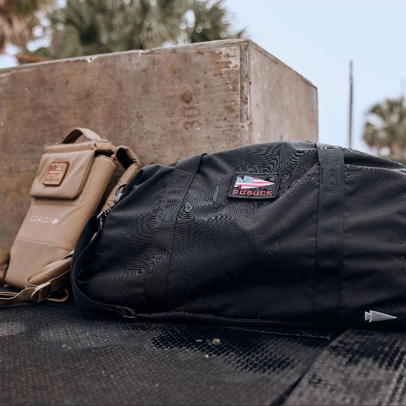 A black duffel bag and tan shoulder bag rest on a surface outdoors near a wooden box and palm trees.