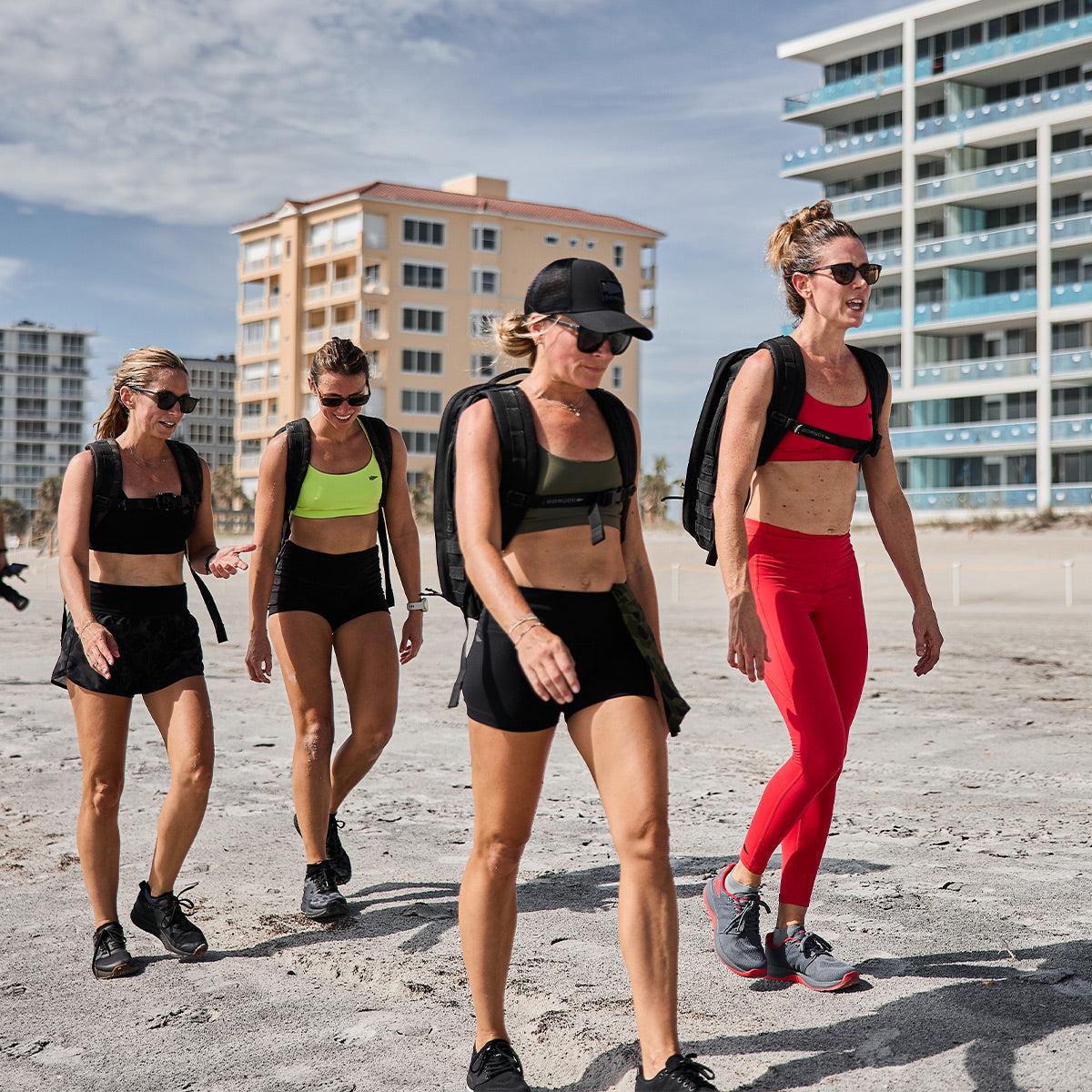 Four women in athletic wear and backpacks walk together on a sandy beach near tall buildings.
