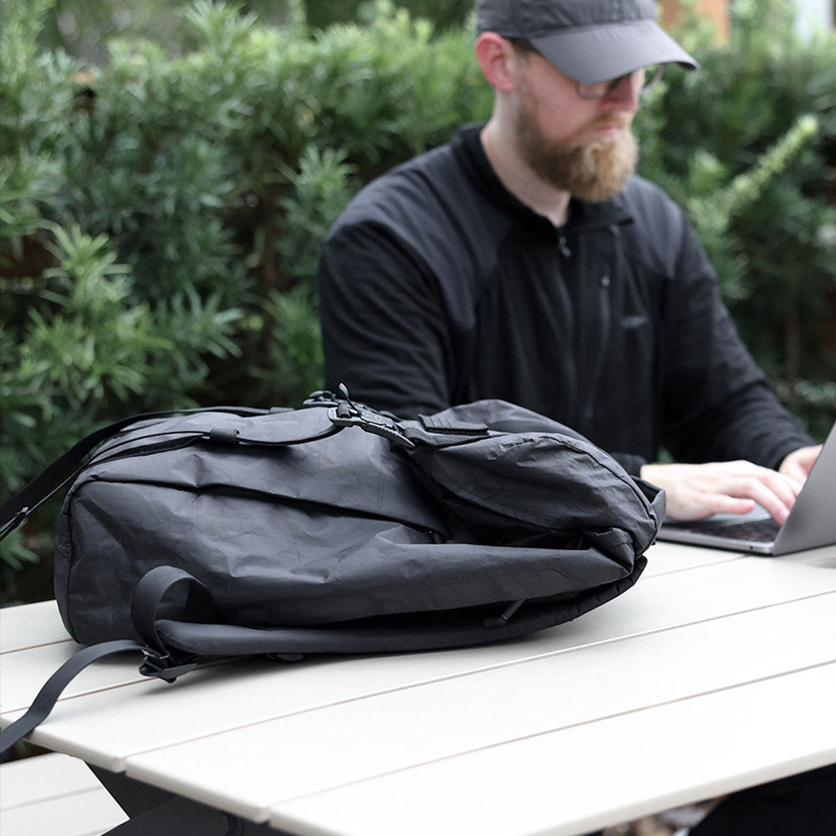 A black backpack on a table with a bearded man working on a laptop in the background outdoors.