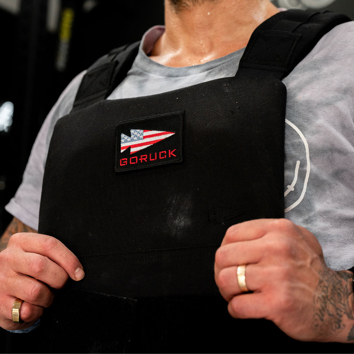 A person in a weighted vest with a GORUCK patch and wedding ring, gripping the vest straps.