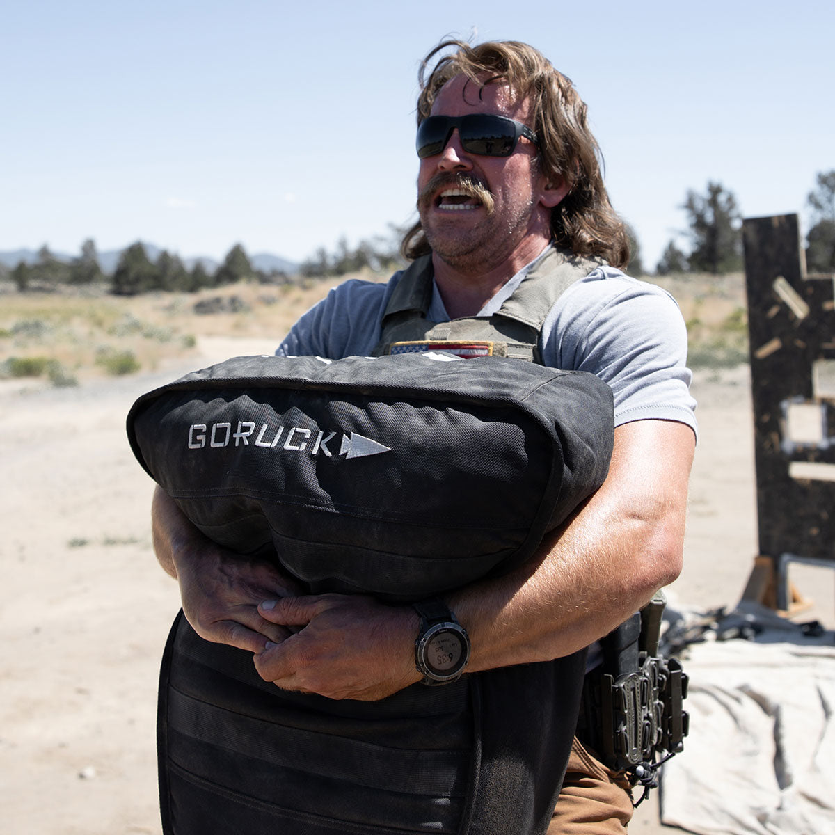 Man outdoors lifting a heavy GORUCK training sandbag, wearing sunglasses and tactical gear in sunny weather.