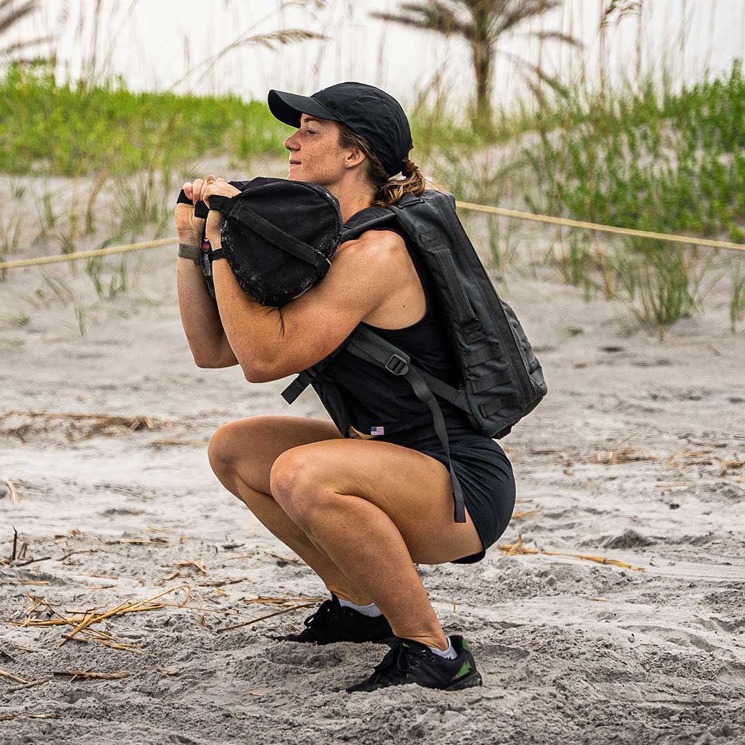 Woman in athletic wear squats on a sandy beach, holding a sandbag with a backpack on her back.