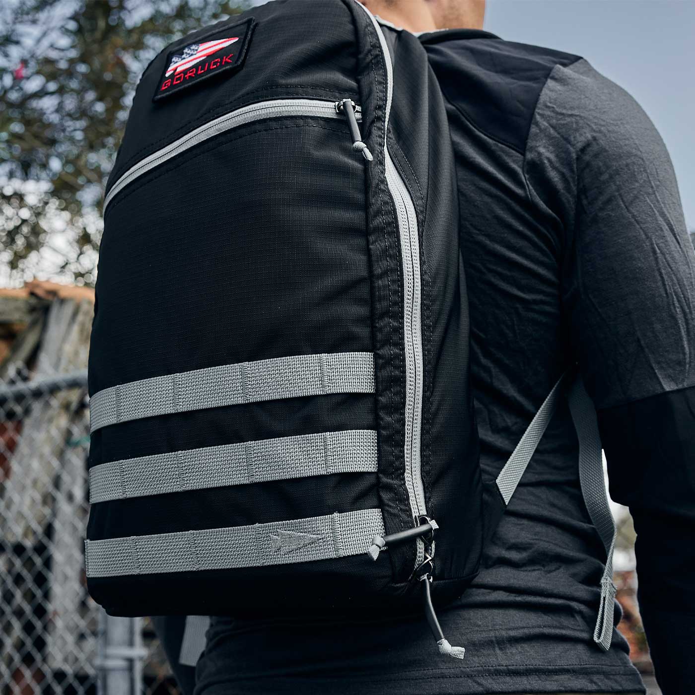 Person wearing a black backpack with gray straps and a red patch, standing outdoors near a fence.