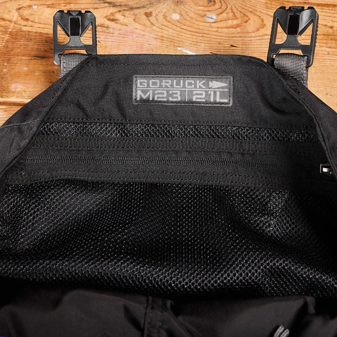 A close-up of a sleek black GORUCK M23 - Cordura backpack laid flat, displaying the label on the upper inside section. The modern rucksack's interior mesh pocket is visible, while the wooden surface beneath adds texture to the image.