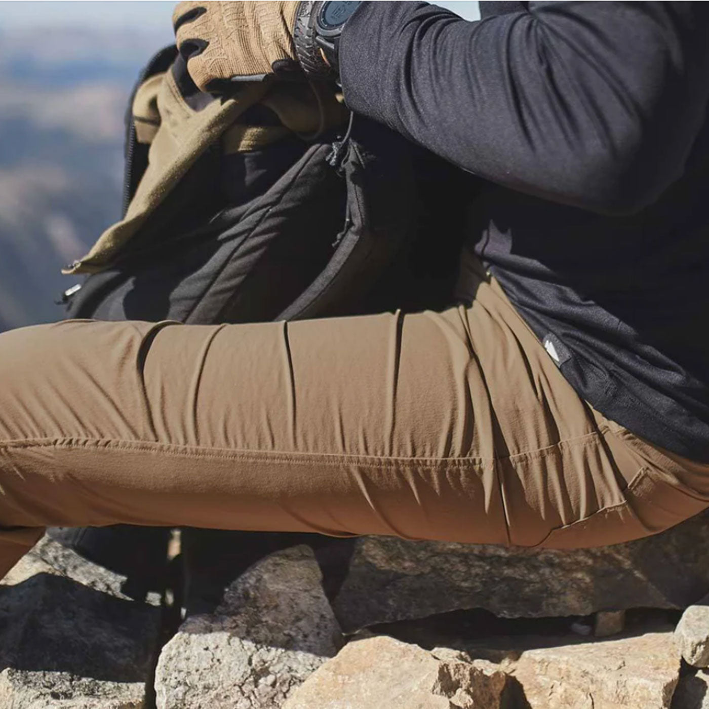 A person in brown pants and gloves sits on rocks with a backpack, outdoors in a mountainous area.