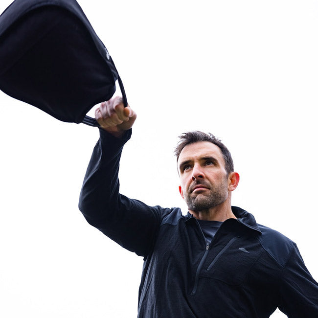 Man in black jacket swings a weighted bag outdoors against a bright, white sky.