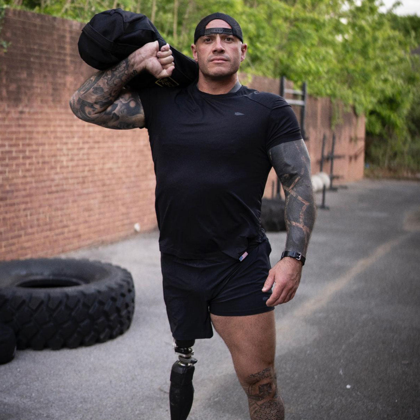 A man with a prosthetic leg carries a heavy bag on his shoulder outdoors near tires and brick wall.