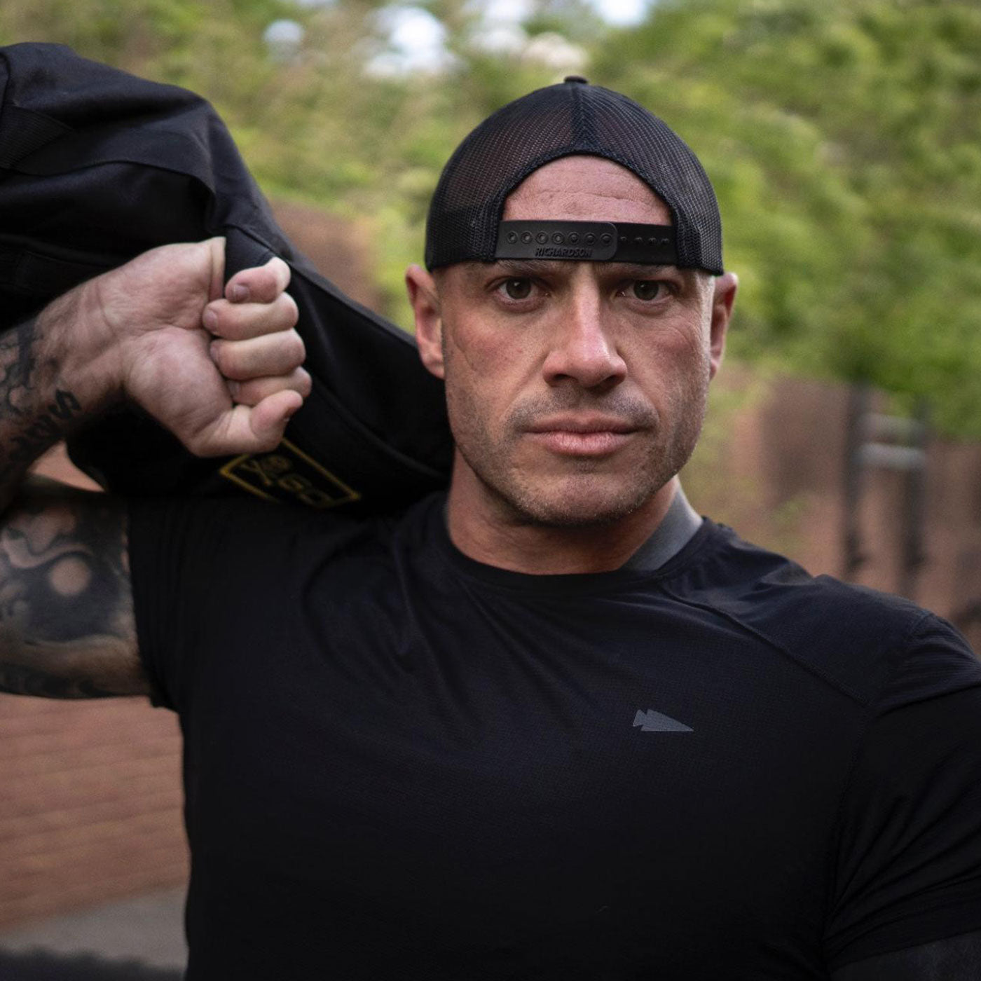 Man in a black shirt and backwards cap holding a sandbag over his shoulder, staring at the camera.
