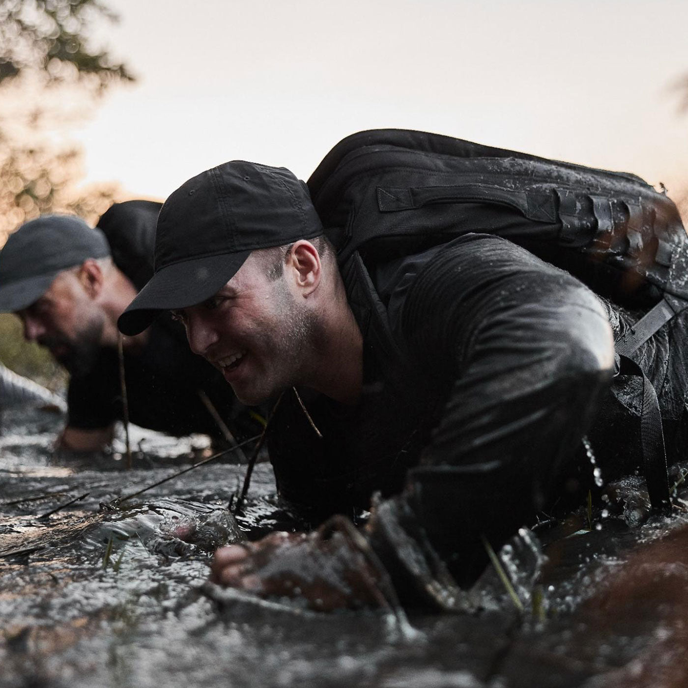 Two people in dark clothing and caps crawl through muddy water outdoors, carrying backpacks.