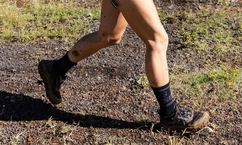 Person wearing hiking boots and black socks walking on a rocky dirt trail with grass in the background.