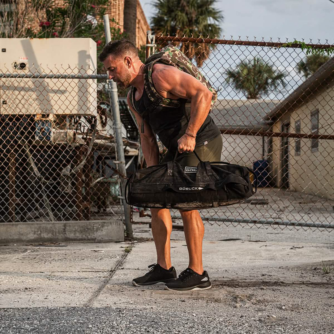 A man lifts a heavy bag outdoors by a chain-link fence, wearing GORUCK Ballistic Trainers in Black + Glacier Grey with Silver Reflective Spearhead and a backpack.