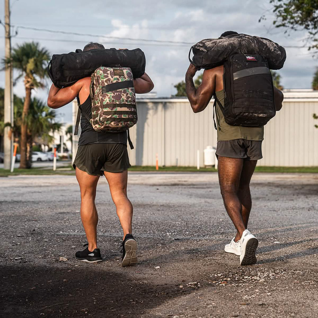 Two men in GORUCK Ballistic Trainers - Black + Glacier Grey with silver reflective spearhead carry backpacks and heavy bags outdoors on a gravel path.