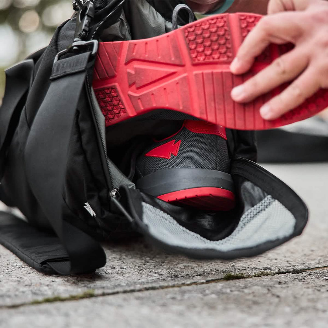 A person packs GORUCK Ballistic Trainers in Wolf Grey + High Risk Red with 3X Stability and a red reflective spearhead into a black gym bag made of durable CORDURA® Ballistic Nylon.