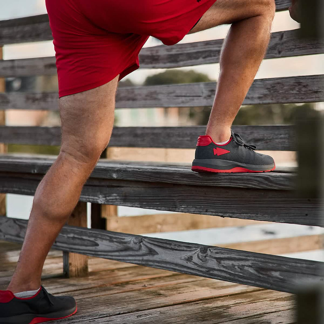 A man in red shorts steps onto a wooden bench outdoors, wearing GORUCK Ballistic Trainers - Wolf Grey + High Risk Red W / Red Reflective Spearhead, designed for functional fitness with 3X Stability.