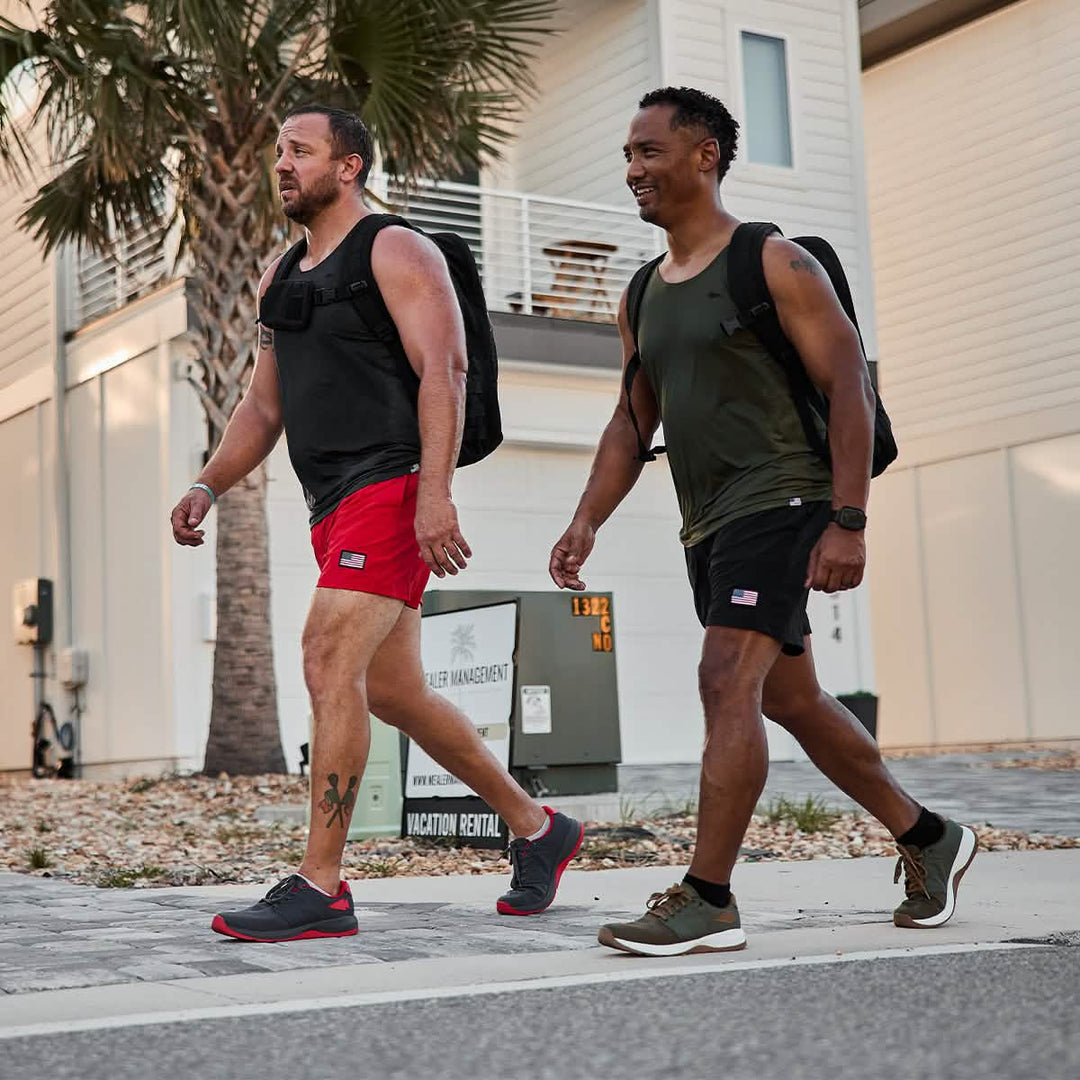 Two men in athletic wear and backpacks walk near palm trees and modern buildings, wearing GORUCK Ballistic Trainers – Wolf Grey + High Risk Red with CORDURA® Ballistic Nylon for 3X Stability on any terrain.