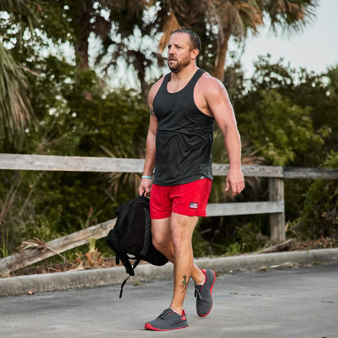 A man in red shorts and a black tank top walks outdoors among trees, carrying the GORUCK Ballistic Trainers - Wolf Grey + High Risk Red W / Red Reflective Spearhead backpack made from durable CORDURA® Ballistic Nylon.