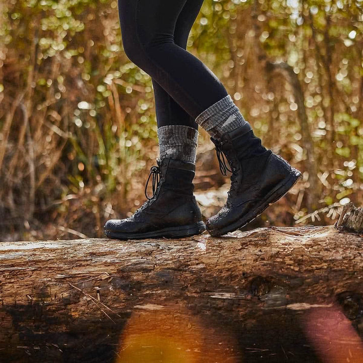 Wearing GORUCK Merino Challenge Socks, a person in black boots and leggings balances on a fallen log in a sunlit forest.