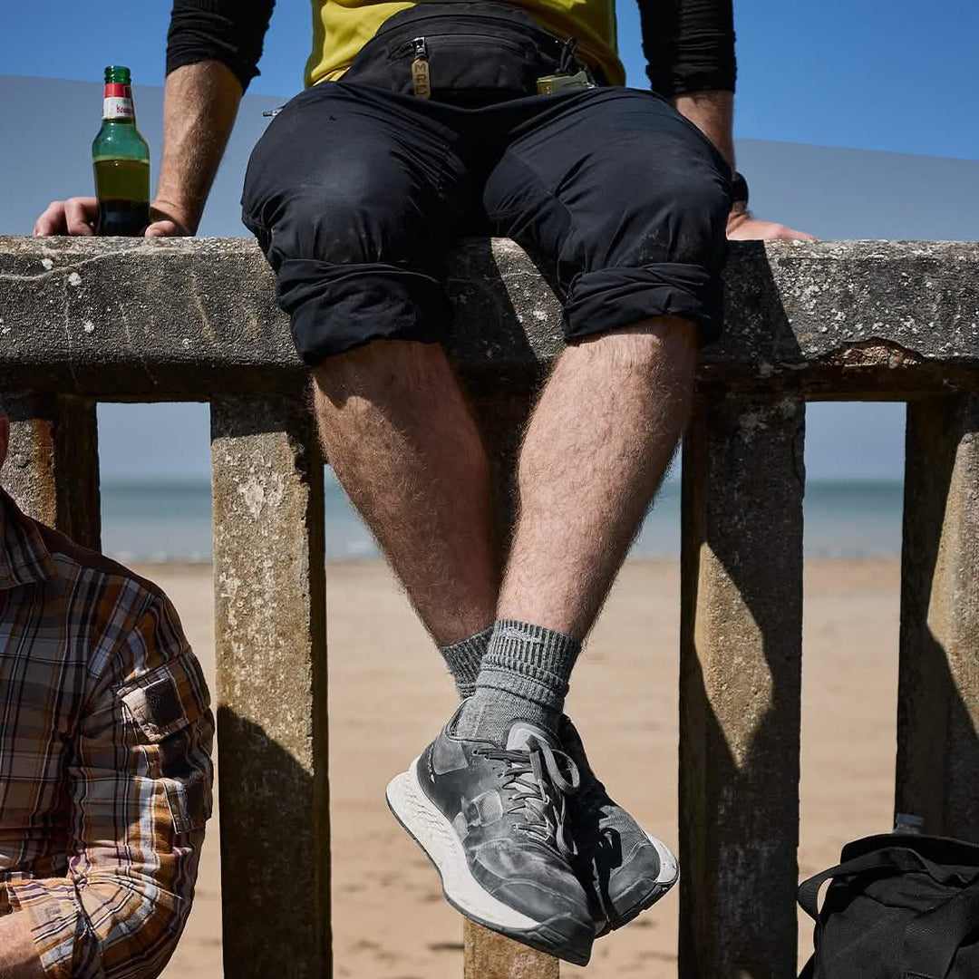 Relaxing on a concrete railing at the beach with crossed legs in GORUCK Merino Challenge Socks, enjoying a beer on a sunny day.